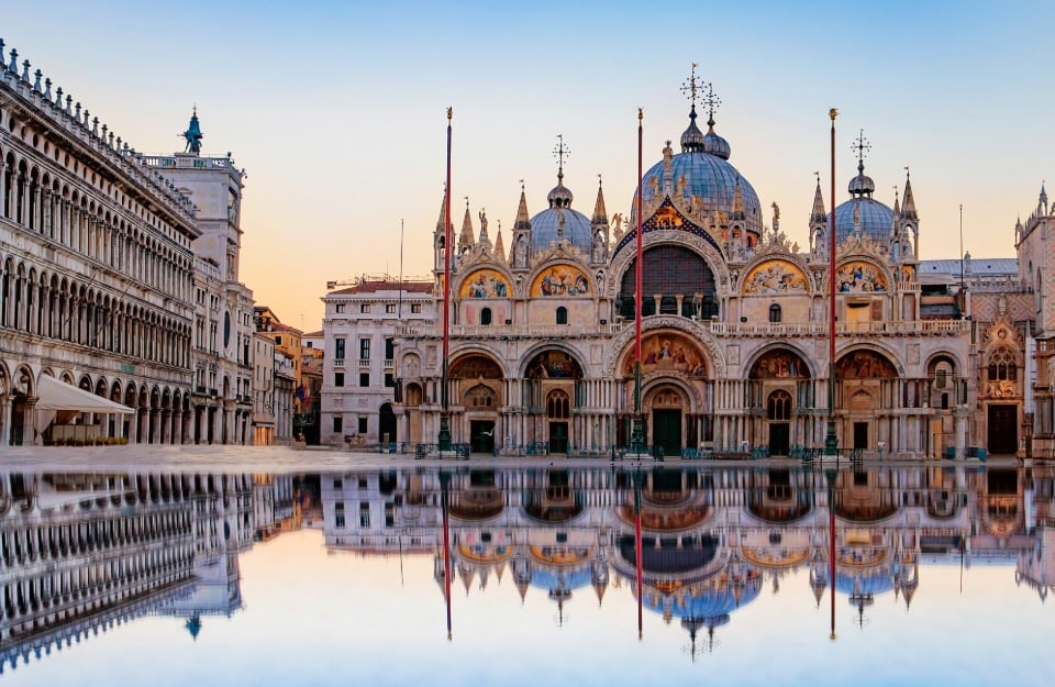 La facciata della Basilica di San Marco a Venezia, Italia, con il suo riflesso in una grande pozzanghera d'acqua in primo piano. La basilica è un famoso esempio di architettura italo-bizantina, caratterizzata dal suo design ornato, dalle cupole e dai mosaici. L'immagine cattura i dettagli intricati dell'esterno della basilica, inclusi gli archi, le colonne e le statue
