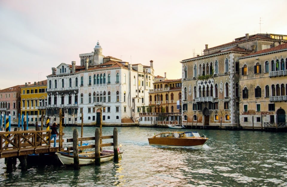 Vista di Venezia, con edifici storici dalle facciate ornate che si affacciano su un canale. Una motolancia di legno naviga sull'acqua, mentre un molo in legno con alcune barche ormeggiate si trova sul lato sinistro. Gli edifici mostrano un mix di stili architettonici, con finestre ad arco e balconi decorati. L'acqua del canale riflette gli edifici e la barca, aggiungendo al fascino della scena. Il cielo sembra essere al tramonto, emanando una luce calda sugli edifici
