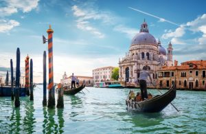 Una scena iconica di Venezia. In primo piano ci sono due gondole con gondolieri che remano attraverso un canale. Sullo sfondo si vede la Basilica di Santa Maria della Salute, un famoso edificio barocco con una grande cupola. L'acqua del canale è verde e riflette il cielo azzurro con alcune nuvole. Ci sono anche pali di ormeggio a strisce arancioni e nere che emergono dall'acqua