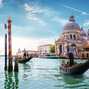 Una scena iconica di Venezia. In primo piano ci sono due gondole con gondolieri che remano attraverso un canale. Sullo sfondo si vede la Basilica di Santa Maria della Salute, un famoso edificio barocco con una grande cupola. L'acqua del canale è verde e riflette il cielo azzurro con alcune nuvole. Ci sono anche pali di ormeggio a strisce arancioni e nere che emergono dall'acqua