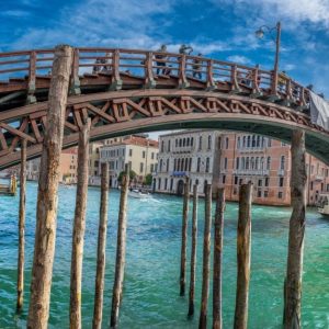 Un ponte di legno che attraversa un canale a Venezia, Italia. Il ponte è costruito con una struttura a traliccio di travi in legno e ha una ringhiera sulla parte superiore. Sotto il ponte, ci sono diversi pali di legno che sporgono dall'acqua. L'acqua del canale è di un vibrante colore turchese e ci sono edifici con la classica architettura veneziana che fiancheggiano il canale. Una barca è visibile sul lato destro dell'immagine e il cielo è sereno con alcune nuvole sparse