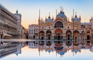 La facciata della Basilica di San Marco a Venezia, Italia, con il suo riflesso in una grande pozzanghera d'acqua in primo piano. La basilica è un famoso esempio di architettura italo-bizantina, caratterizzata dal suo design ornato, dalle cupole e dai mosaici. L'immagine cattura i dettagli intricati dell'esterno della basilica, inclusi gli archi, le colonne e le statue