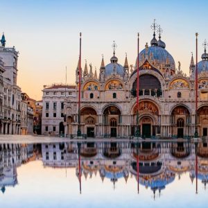 La facciata della Basilica di San Marco a Venezia, Italia, con il suo riflesso in una grande pozzanghera d'acqua in primo piano. La basilica è un famoso esempio di architettura italo-bizantina, caratterizzata dal suo design ornato, dalle cupole e dai mosaici. L'immagine cattura i dettagli intricati dell'esterno della basilica, inclusi gli archi, le colonne e le statue