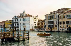 Vista di Venezia, con edifici storici dalle facciate ornate che si affacciano su un canale. Una motolancia di legno naviga sull'acqua, mentre un molo in legno con alcune barche ormeggiate si trova sul lato sinistro. Gli edifici mostrano un mix di stili architettonici, con finestre ad arco e balconi decorati. L'acqua del canale riflette gli edifici e la barca, aggiungendo al fascino della scena. Il cielo sembra essere al tramonto, emanando una luce calda sugli edifici