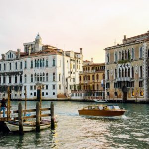 Vista di Venezia, con edifici storici dalle facciate ornate che si affacciano su un canale. Una motolancia di legno naviga sull'acqua, mentre un molo in legno con alcune barche ormeggiate si trova sul lato sinistro. Gli edifici mostrano un mix di stili architettonici, con finestre ad arco e balconi decorati. L'acqua del canale riflette gli edifici e la barca, aggiungendo al fascino della scena. Il cielo sembra essere al tramonto, emanando una luce calda sugli edifici